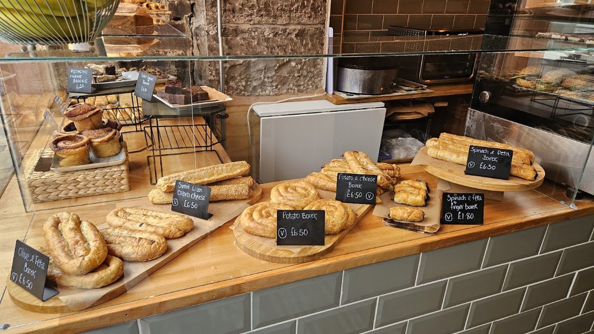 The glass counter: coiled and long börek on wooden boards, sesame simit, and trays of spinach and feta fingers, each with a handwritten chalk label.