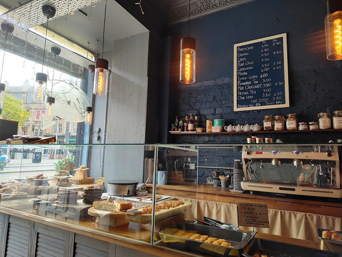Inside Kukina: amber filament lamps hanging over a glass counter of pastries, a navy-painted brick wall with a chalk drinks board and a shelf of teapots and jars, and the espresso machine behind.