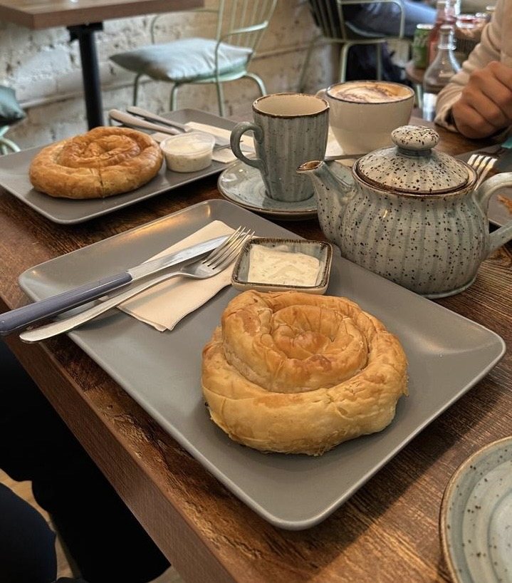 A round coiled börek on a grey plate with a dish of yoghurt, beside a speckled teapot, a cup and a cappuccino.