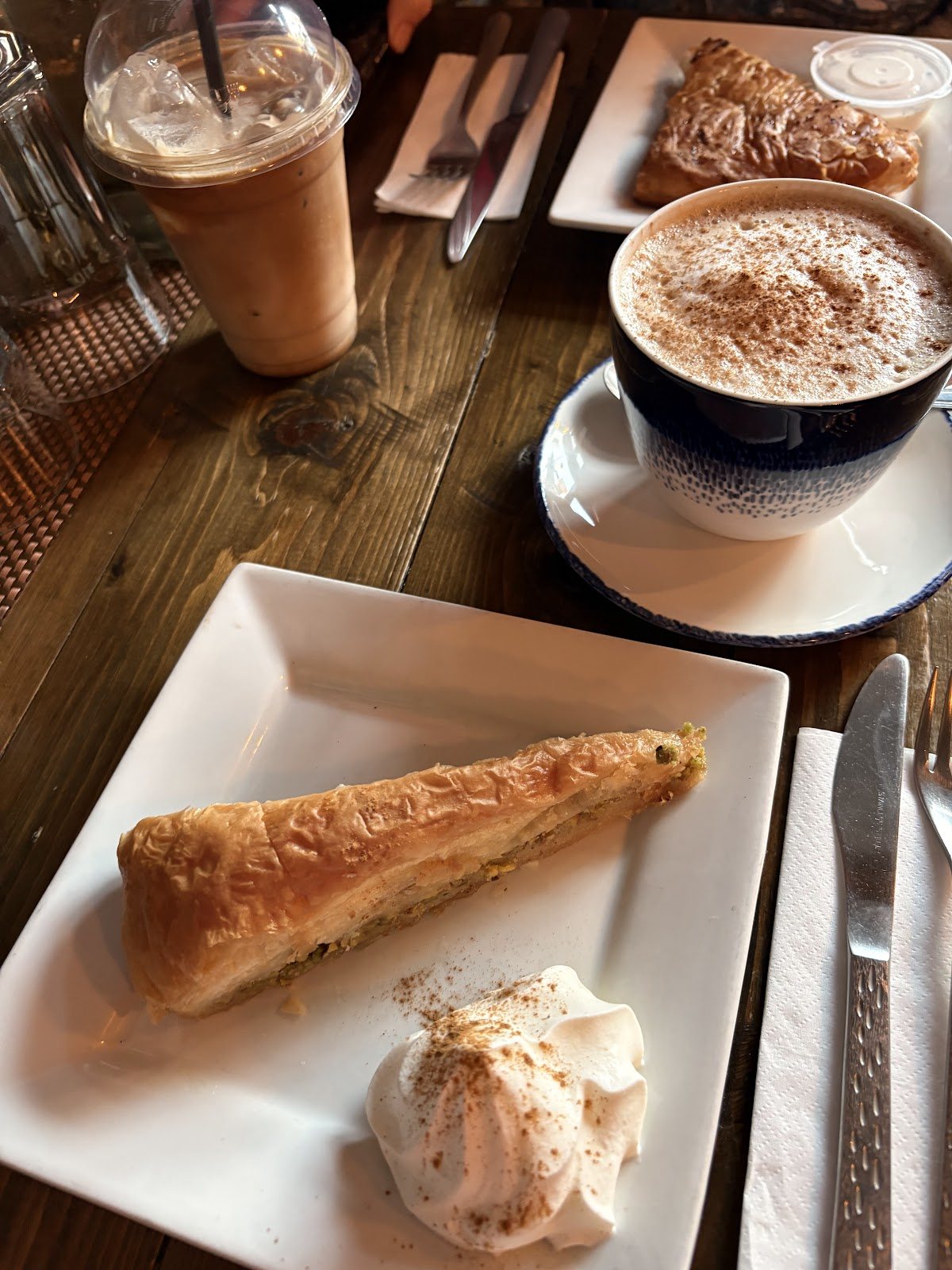 A wedge of layered pastry with whipped cream and a dusting of cinnamon, beside a large cinnamon-topped cappuccino and an iced coffee.